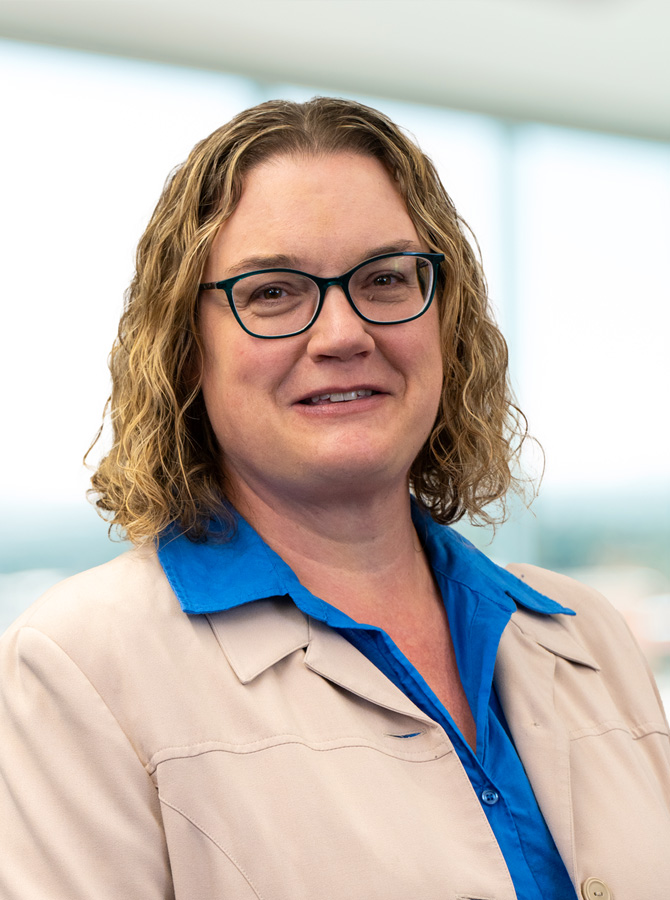 Woman with short curly blonde hair with glasses wearing a tan blazer over a royal blue collared shirt