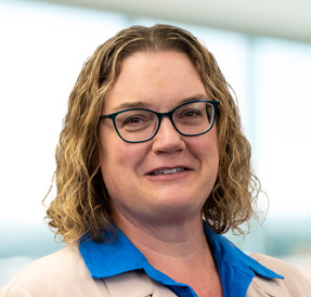 Woman with short curly blonde hair with glasses wearing a tan blazer over a royal blue collared shirt