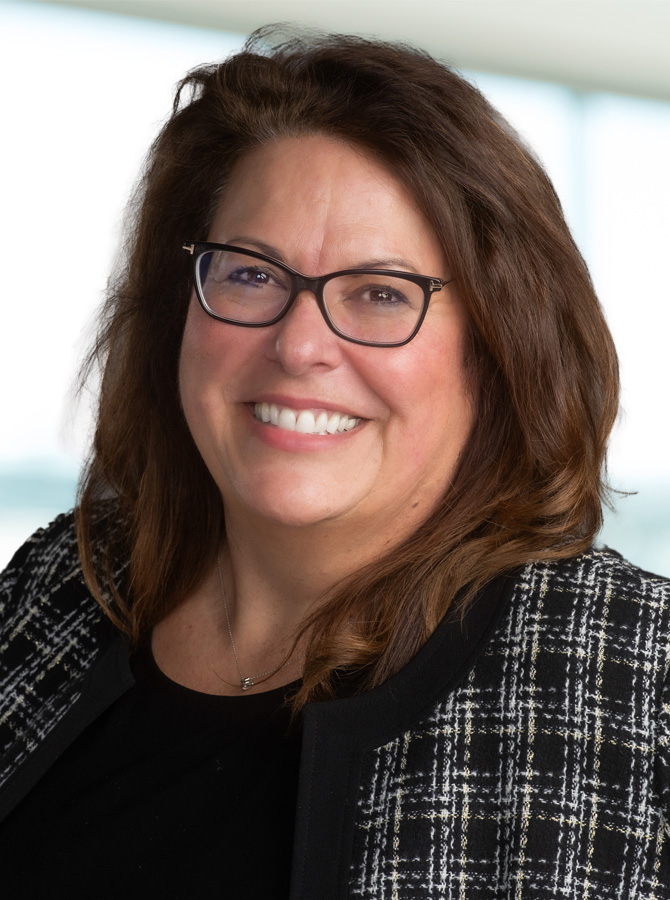 Light skinned woman with medium brown hair in a black jacket wearing black-rimmed glasses, smiling with teeth showing