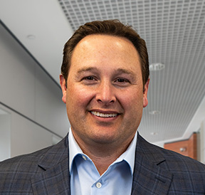 light skinned man named Mike with short brown hair wearing a light blue shirt and charcoal grey jacket.